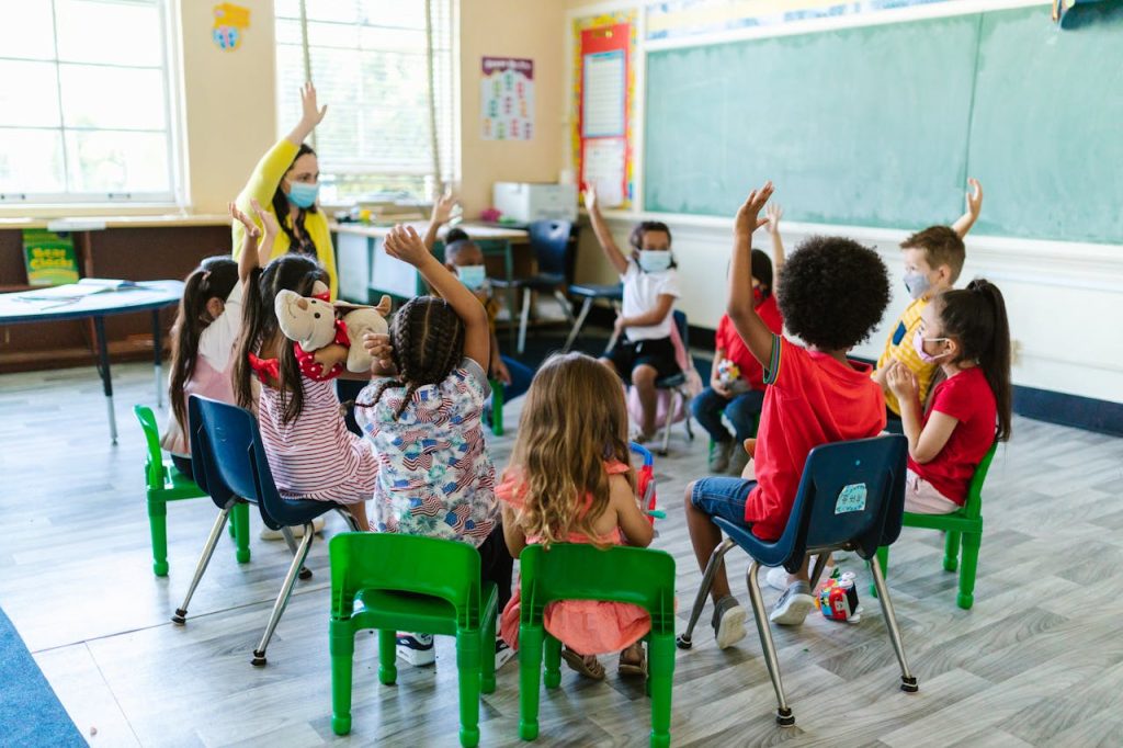 people-sitting-on-chairs-inside-the-room-8364026 Diverse group of children in a classroom, raising hands, led by masked teacher. Bright and engaging learning environment.